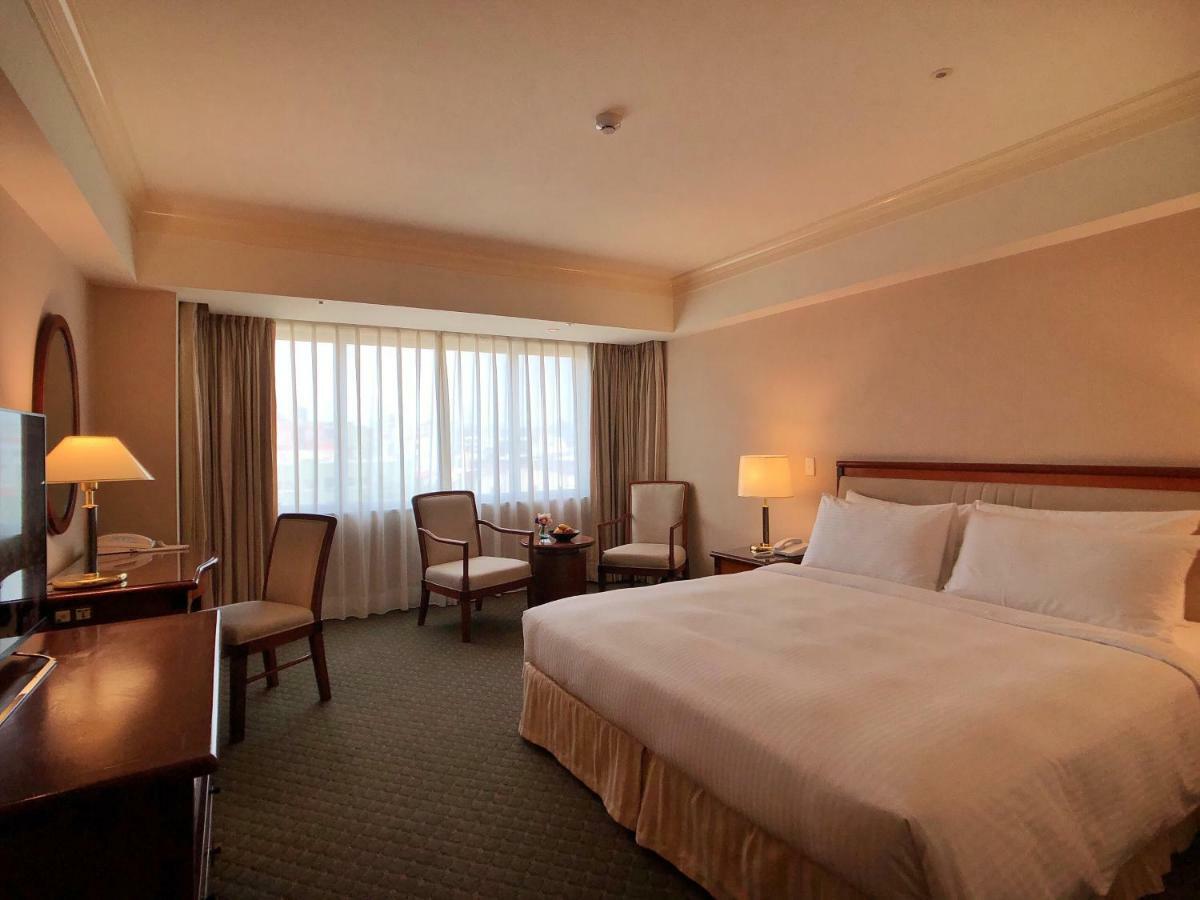 เอเวอร์กรีน เลาเรล โฮเต็ล ไทชุง Hotel ไถจง ภายนอก รูปภาพ The photo shows a hotel room featuring a large bed with white linens, a wooden bedside table with a lamp, and a flat-screen television. There are two chairs with a small table next to them positioned by a large window, which has sheer curtains lettin